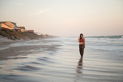 High school senior girl in harem pants and crochet crop top walking along water soaked beach at sunset