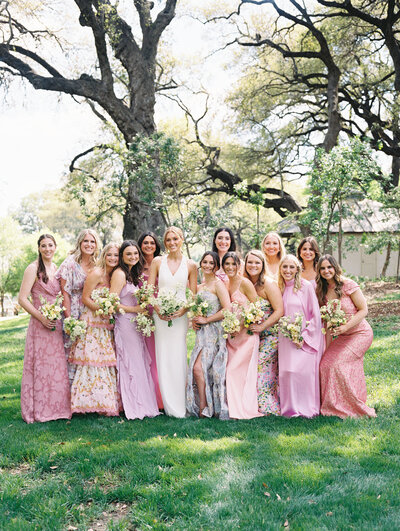 Bride surrounded by bridesmaids in different pink dresses in front of a large tree