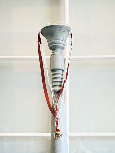 A red ribbon with a small gold bell, crafted as a charming holiday ornament, hangs from the top of a white painted wooden post in front of a window.