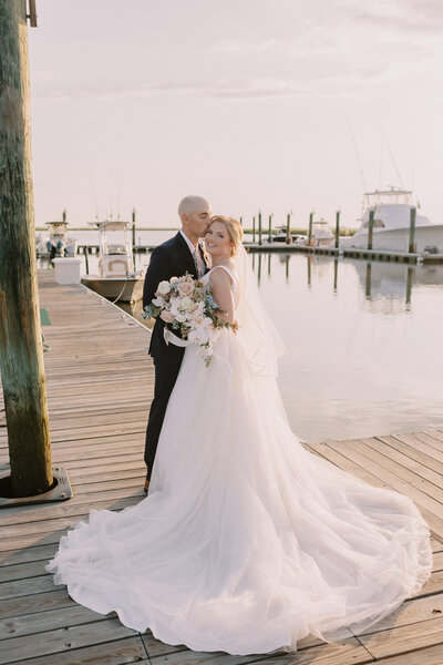 Groom in black tux kisses bride on cheek who is looking over her shoulder at camera. She is holding a summer spray bouquet of pastel flowers and they are standing on a boat dock with white fishing boats in the background.