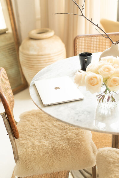 An interior scene with a table set with cream roses, a black coffee cup and a closed macbook pro