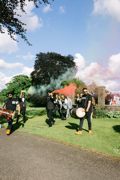 Candid image of the groom on the way to get married he is walking with his friends who are lighting smoke bombs followed by drummers as he walks to the ceremony at Luttrellstown Castle
