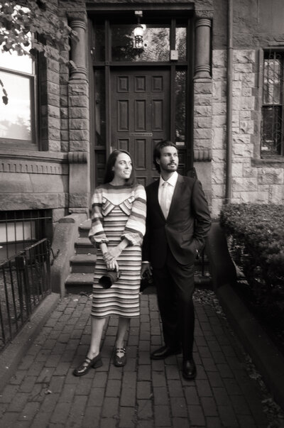 A black-and-white 35mm film photograph capturing a wedding photography duo during a branding session in Boston. The pair stands confidently in front of a historic brownstone, exuding professionalism and charm. One holds a camera, symbolizing their craft, while their stylish attire reflects a blend of elegance and approachability, perfectly aligning with their wedding photography brand.