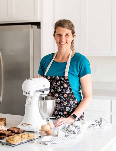 Branding photo of Jenny's Cookie Company with the baker standing in her kitchen with ther Kitchenaid and baking ingredients smiles at the camera for her headshot with  Colorado branding photographer Avenir Photo Co.