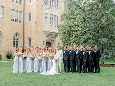 Wedding party in black tuxes and light blue dresses standing in a line smiling forward with the University of St. Thomas campus building behind them.