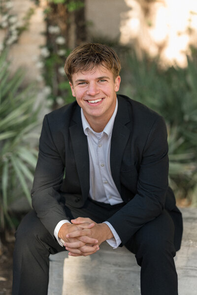 San Antonio senior guy in a suit leans forward on his knees and smiles at the camera.