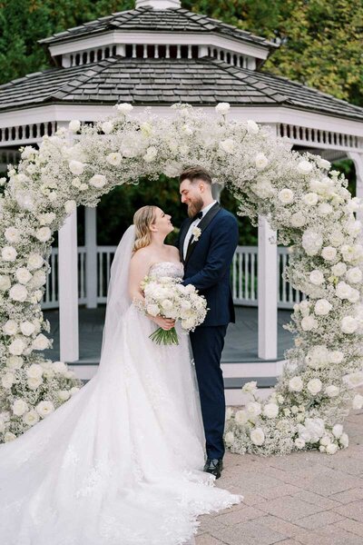 Bride and groom standing under a white floral arch at Deer Creek in Ajax, photographed by Ontario wedding photographer Nadtochiy