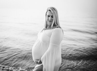 Pregnant woman in a white dress by the water, captured in black and white