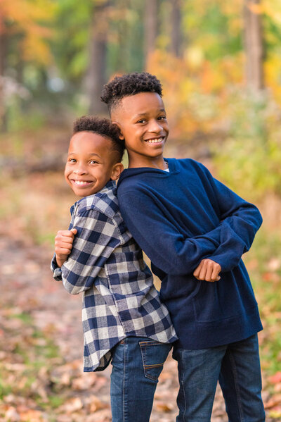 Two young dark-skinned brothers back to back in the fall woods; photo by Chicago family photographer Kristen Hazelton