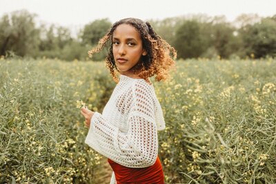 Woman with curly brown and blonde hair looks over her shoulder in a field of small yellow flowers, holding a few blossoms.