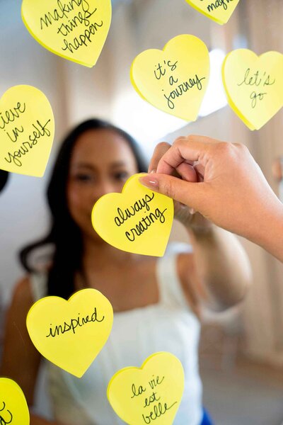 Creative brand photographer engaging with a motivational quote on a yellow sticky note, placing it on a mirror that reflects her brand identity