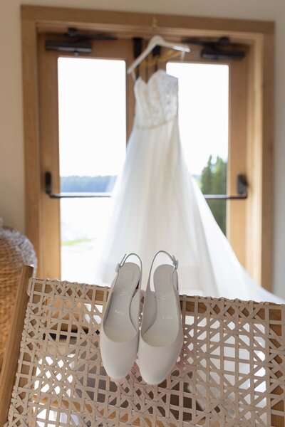 Rhode Island Wedding Photographer | Kelsey Sheehan Photography Timeless Rhode Island Weddings | White bridal shoes rest on a wicker chair in front of a window, where a flowing wedding dress hangs elegantly. Bright, serene atmosphere.