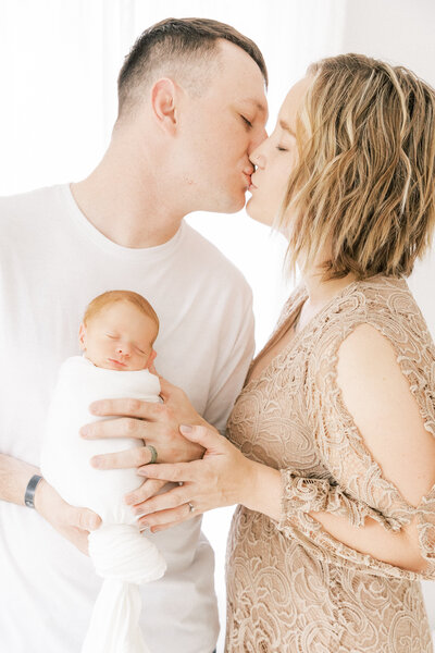 Mom and dad kissing while holding their preemie newborn baby girl during studio photoshoot