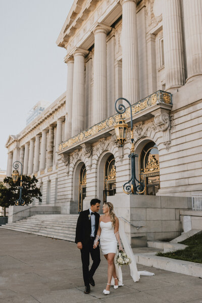 A newlywed couple walking hand in hand outside San Francisco City Hall with large columns and intricate golden decorations. the bride wears a short white dress and holds a bouquet.