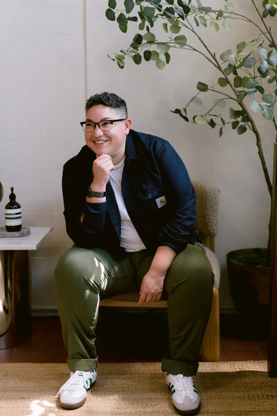 Confident individual with short hair and glasses, seated in a modern, light-filled space with greenery, wearing a casual yet polished outfit.