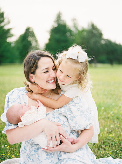 Mother sits in field holding newborn baby daughter as her toddler wraps her up from behind and they both giggle