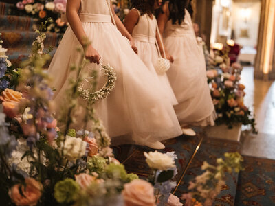 A charming moment at Ashridge House in Hertfordshire, as little flower girls make their way down the grand staircase before the bridal walk. The image captures their delicate dresses flowing over each step, tiny feet in dainty shoes, and a small hand gently holding a flower crown, pure elegance and anticipation in every detail.