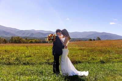 A bride and groom stand facing each other embracing with their foreheads together and arms wrapped around each other. The bride is in a fitted wedding gown and the groom is in a black suit. The are standing in a green grassy field with the sun shining down on them and a vast mountain range in the background.