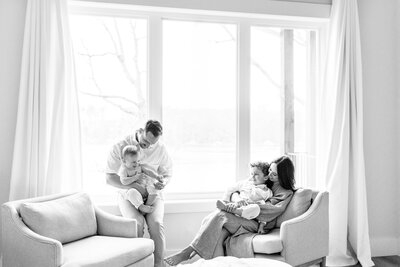 Black and white image of a Kansas City family with two young children sitting together by a large window during their lifestyle photography session.