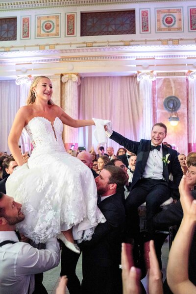A bride and groom are joyfully lifted up on chairs by wedding guests in a lavish ballroom. The bride wears a white gown, and the groom is in a suit. The room is elegantly decorated, filled with smiling guests celebrating.