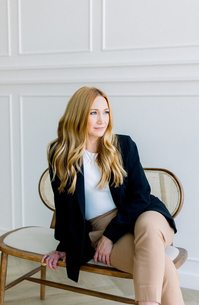 Redheaded woman in black blazer sitting on a rattan chair and looking away from camera in Calgary, AB.