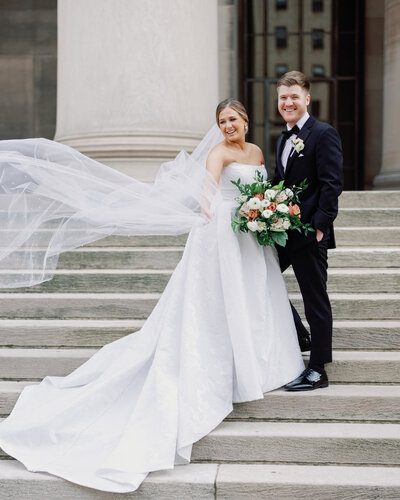 A bride’s veil dances in the air as she smiles with her groom during golden hour portraits outside a historic Pittsburgh location.