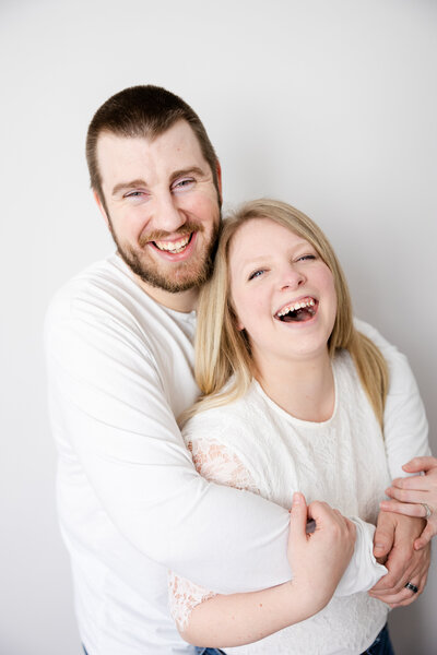 Husband and wife snuggling close at home in Beavercreek, Ohio while laughing at the camera.