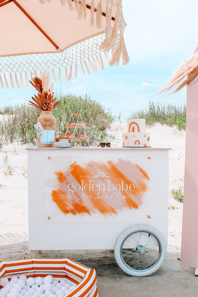 The 'Le Bar Cart' by Shindig Social, featuring a white bar cart with orange brushstroke accents and a sign reading 'golden babe.' The cart is set up on a beach with drinks, a parasol, and beach decor, creating a chic and summery setting.