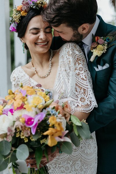 A close-up of a smiling bride and groom. The bride, wearing a flower crown and holding a colorful bouquet, is embraced by the groom, who has a peacock feather boutonniere.