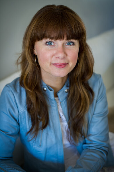 Headshot of marketing strategist Leona Laurie sitting on white couch wearing blue leather jacket.