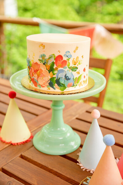 sweet heather ann garden party styled shoot with painted flower cake on green vintage cake stand surrounded by classic birthday party hats