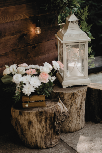 At the entrance of the venue, a charming display of wedding decor welcomes guests with an enchanting ambiance. Lush floral arrangements, bursting with vibrant blooms in shades of blush pink, ivory, and soft greens, adorn rustic wooden barrels placed strategically around the entrance. Interspersed among the florals are elegant lanterns, their warm candlelight casting a soft glow on the surroundings. The flickering light creates a magical atmosphere, illuminating the beauty of the flowers and adding a touch of romance to the scene. Against the backdrop of the venue's facade and the evening sky, this enchanting display sets the tone for a memorable celebration filled with love and joy.