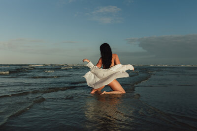 A woman kneels in shallow ocean water at sunset with her wet hair down and white shirt flowing as waves roll in behind her.