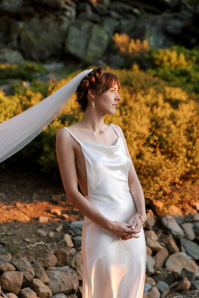 California film photographer as a bride herself, standing in the last bit of light before sunset, looking off to the right. The bride has her hair in a braid and is wearing a veil. She has her hands together in front of her, and she is wearing a white silk dress. She is standing in front of a rock formation with shrubs that look yellow in the sunlight