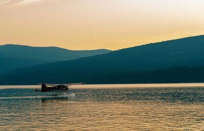 Silhouette of a floatplane landing on a peaceful lake at sunset, with layered mountain ridges in the background, captured by Joseph Mayden for his fine art landscape collection.