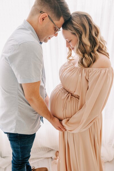 pregnant woman and her husband standing in front of white curtains over a window. they are both looking down and smiling at her pregnant belly.