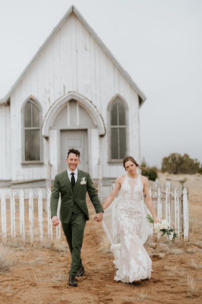 bride and groom holding hands walking with little white chapel