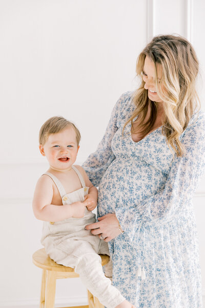 Mother holding toddler son, both wearing light dresses, standing in front of a white wall