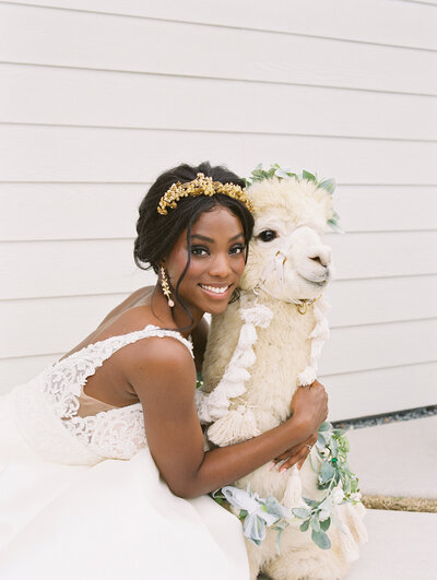 An African-American bride wearing a sleeveless lace wedding gown, Erin Rhyne Headband, and gold drop earrings hugs a llama wearing a flower necklace on the outdoor porch of Boxwood Manor