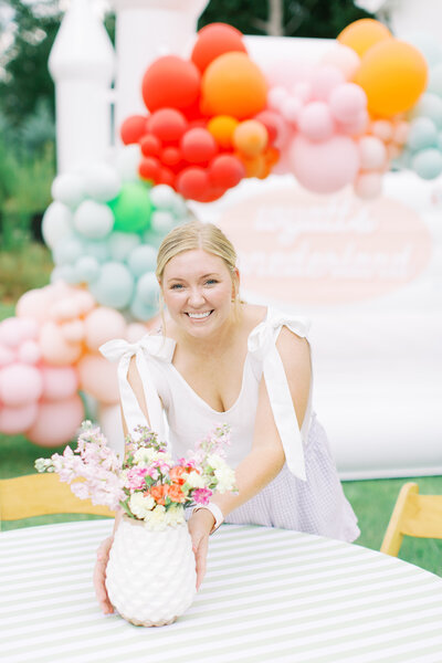 Blonde woman placing vase with flowers on table with white bounce house and colorful balloon arch in background