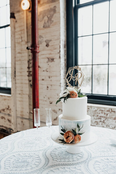White wedding cake with orange flowers and golden initials on top in front of industrial window and exposed brick