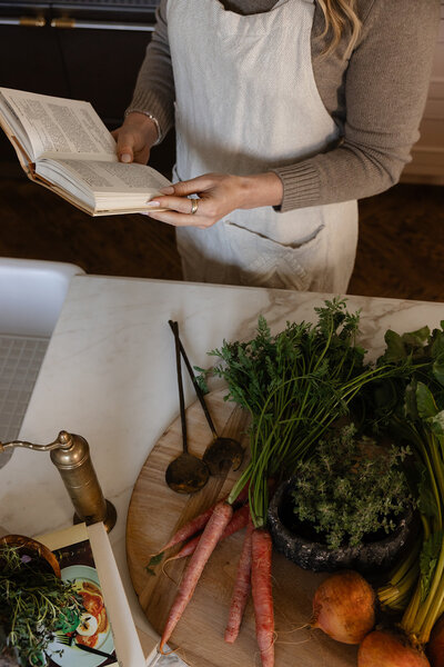 Hands of functional nutritionist Tracy Greenstein holding a book with fresh vegetables and herbs being prepared on the kitchen counter