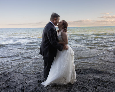A couple is photographed during their elopement while canoeing. They are in a red canoe, with the bride up front and the groom in the back, rowing. She is in a white dress and he is in a brown suit.