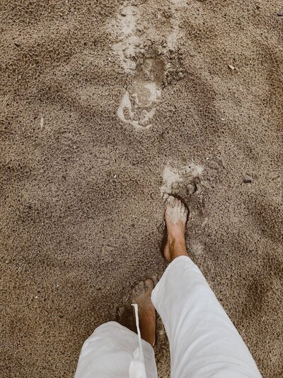 A photograph looking down of a woman’s feet walking through sand in white linen trousers.
