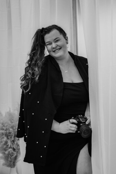 Black and white portrait of a professional photographer dressed in dark clothing, holding a camera up to their face to take a photo, posed against a bright minimalist background—ideal branding image for creative professionals or wedding photographers. 