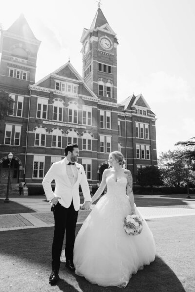 A newlywed couple walks hand in hand in front of Auburn University’s iconic Samford Hall, their smiles radiating pure joy. The groom, dressed in a white tuxedo, and the bride, in a stunning ballgown, share a timeless moment captured in elegant black and white. This breathtaking image showcases the artistry of an Auburn, Alabama wedding photographer, preserving the magic of a classic Southern wedding.
