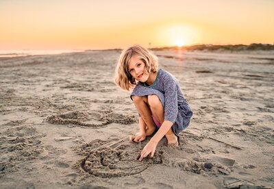 Young girl crouching to draw a rainbow in the sand while smiling during a beach session