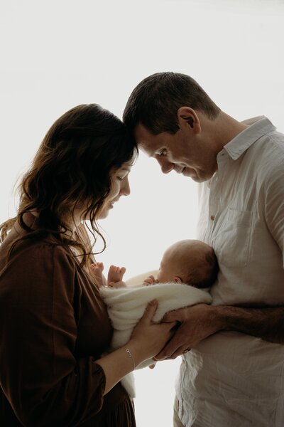 A couple tenderly hold their swaddled newborn, foreheads touching and smiling lovingly at the baby.