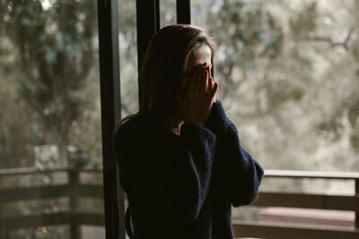 Person inside in front of windows with hands covering face, Struggling with chronic illness ;feeling alone