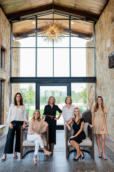 Portrait of six women posing, some standing up and some sitting in chairs in front of a large wall of windows